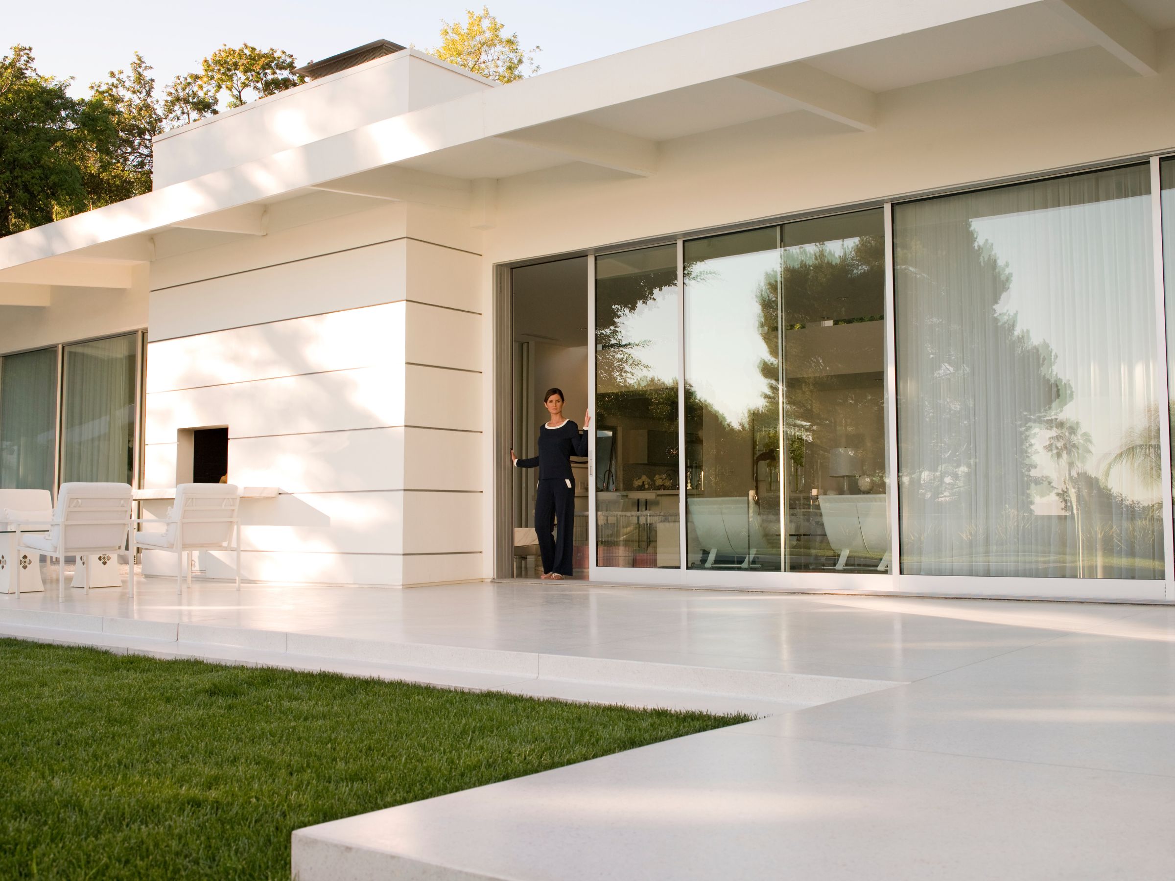 A lady looking out into the back yard of a nice, white house in southern ca with grass and a patio.